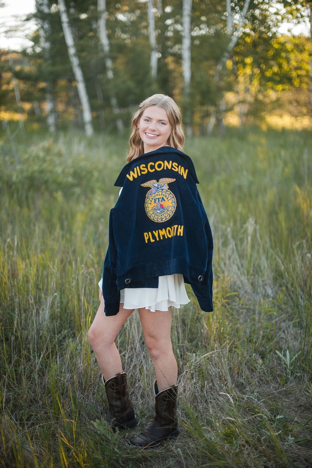 Smiling teenage girl wearing Wisconsin Plymouth FFA jacket, white skirt, and cowboy boots standing in a green field.