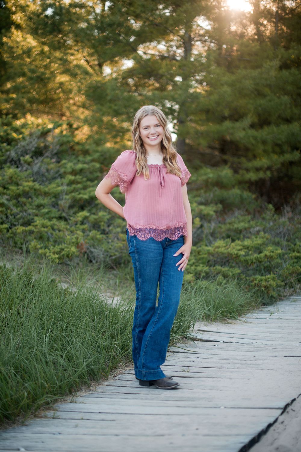 A smiling teenage girl in a pink top and jeans stands on a wooden boardwalk surrounded by lush green trees.