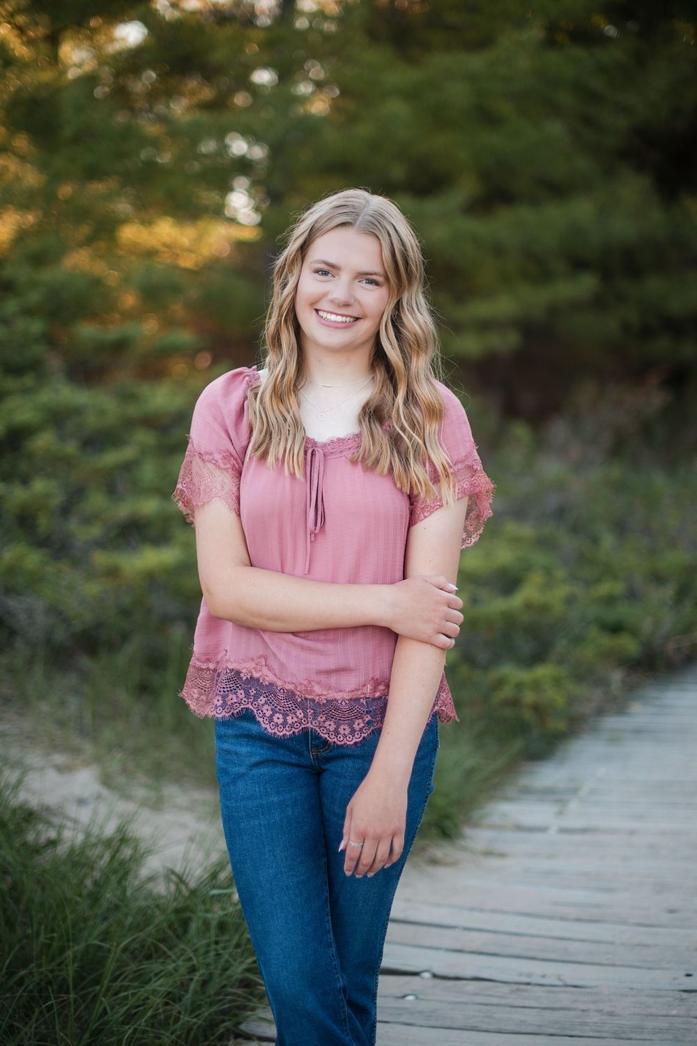 Smiling young woman in pink lace top and jeans posing on a wooden boardwalk surrounded by lush greenery.