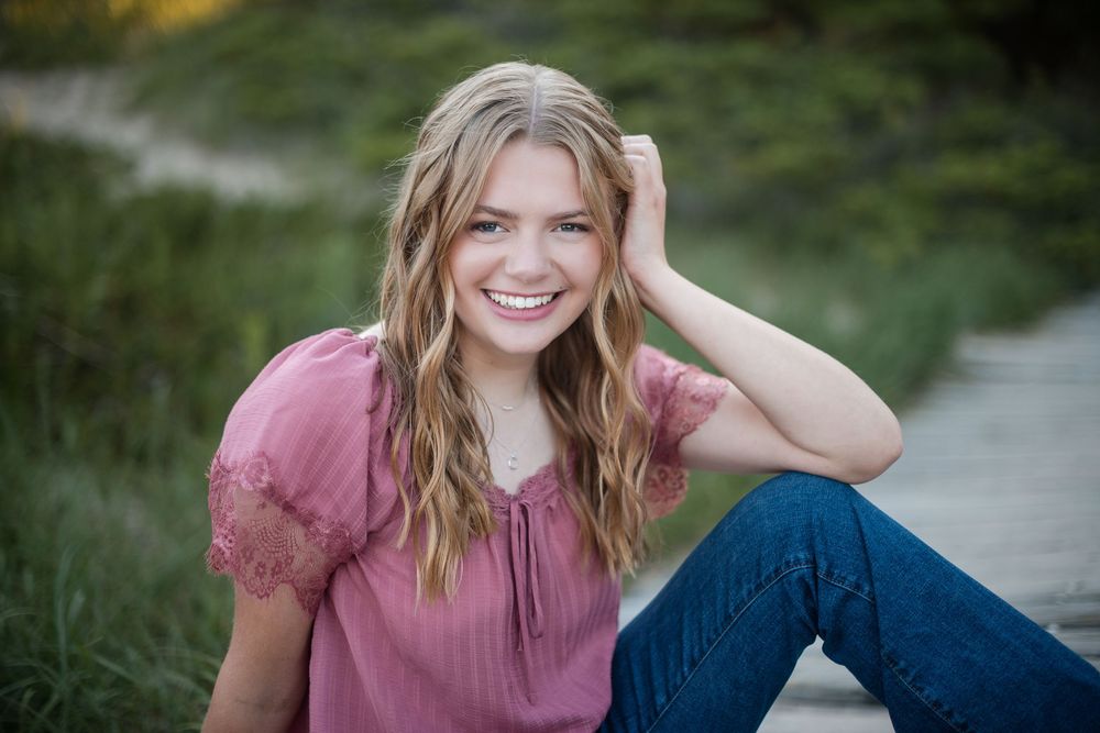 Smiling teenage girl with wavy blonde hair in pink blouse and jeans sitting outdoors for senior portrait.