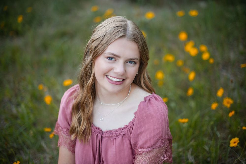 Smiling teenage girl in pink dress sitting in a field of yellow wildflowers for senior portrait photography.