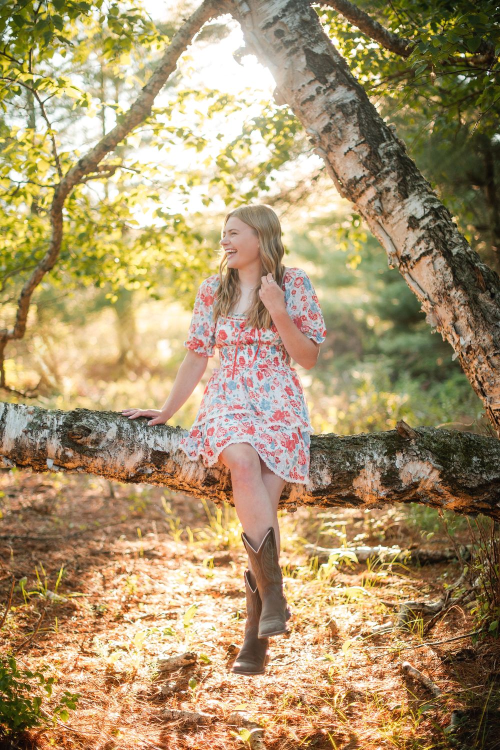 Young woman in floral dress and cowboy boots leaning on birch tree in sunlit forest.