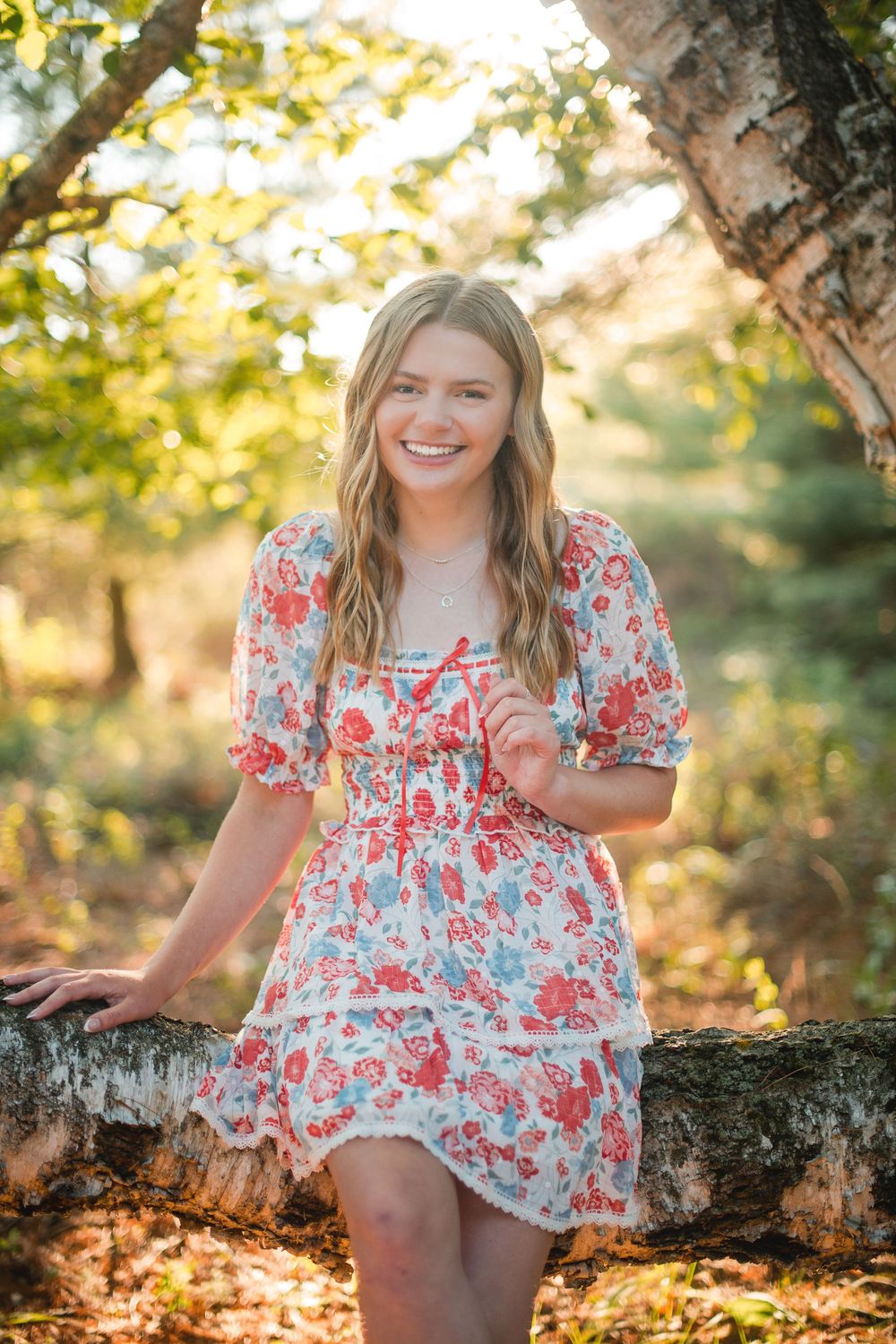 Smiling teenage girl in floral dress leaning on birch tree in sunlit forest during golden hour.