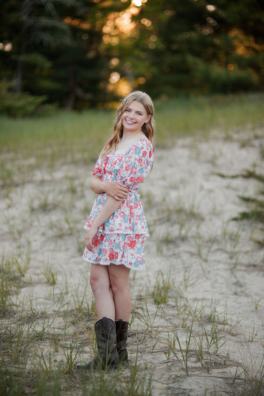 Young woman in floral dress and cowboy boots posing on sandy path with pine trees at sunset.
