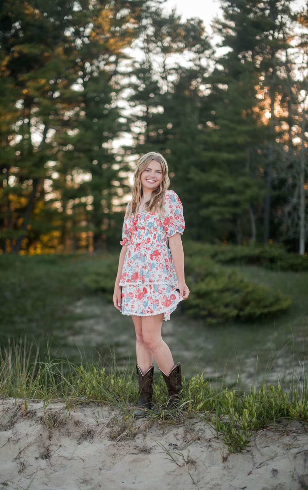 Young woman in floral dress and cowboy boots smiling outdoors on sandy path surrounded by trees at sunset.