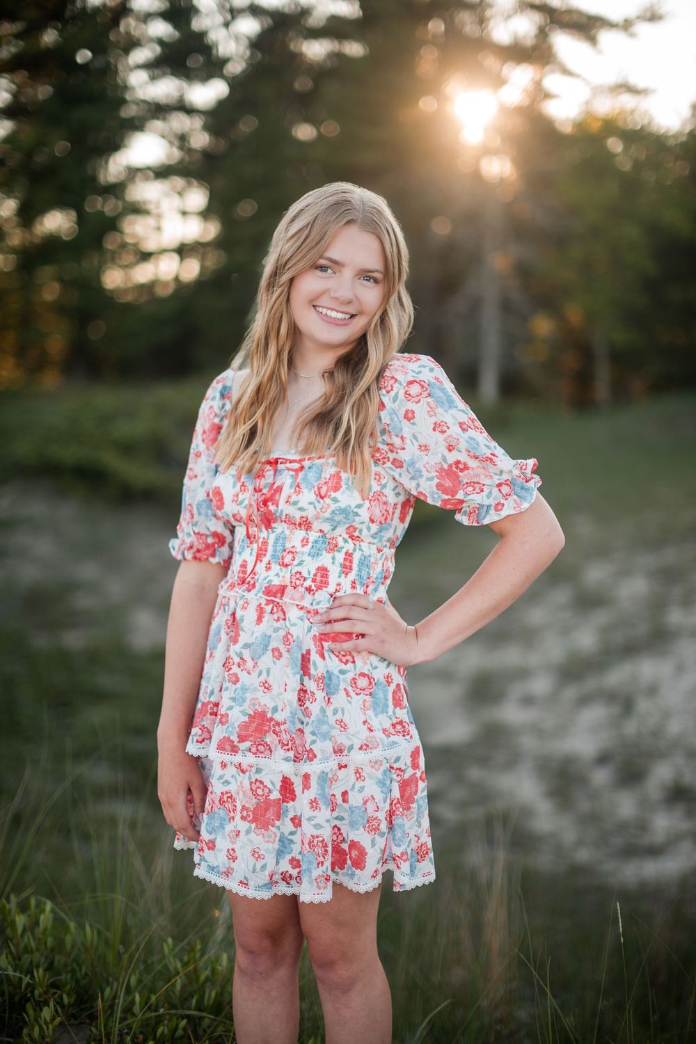 Smiling teenage girl in floral dress poses outdoors at golden hour with sunlit trees in background.