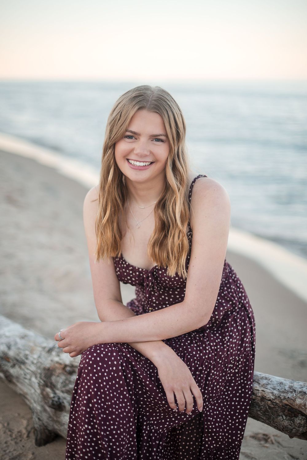 Smiling young woman in purple polka dot dress sitting on driftwood at a sandy beach with ocean in background.