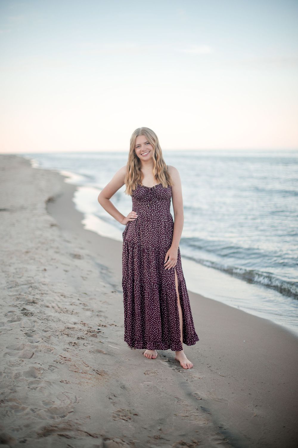 Young blonde woman in floral maxi dress posing barefoot on sandy beach at sunset with ocean waves behind her.