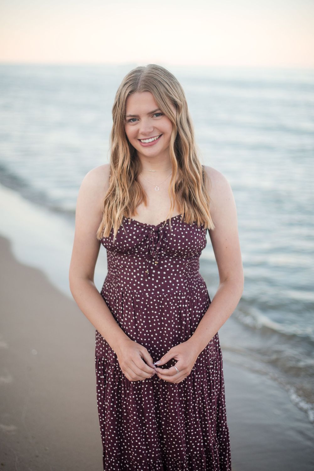 Smiling young woman in purple polka dot sundress standing on a sandy beach with ocean waves behind her.
