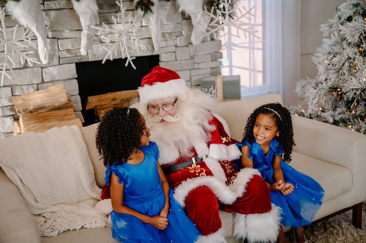 Family of four poses by a flocked Christmas tree, parents and two girls in matching blue dresses and formal wear.