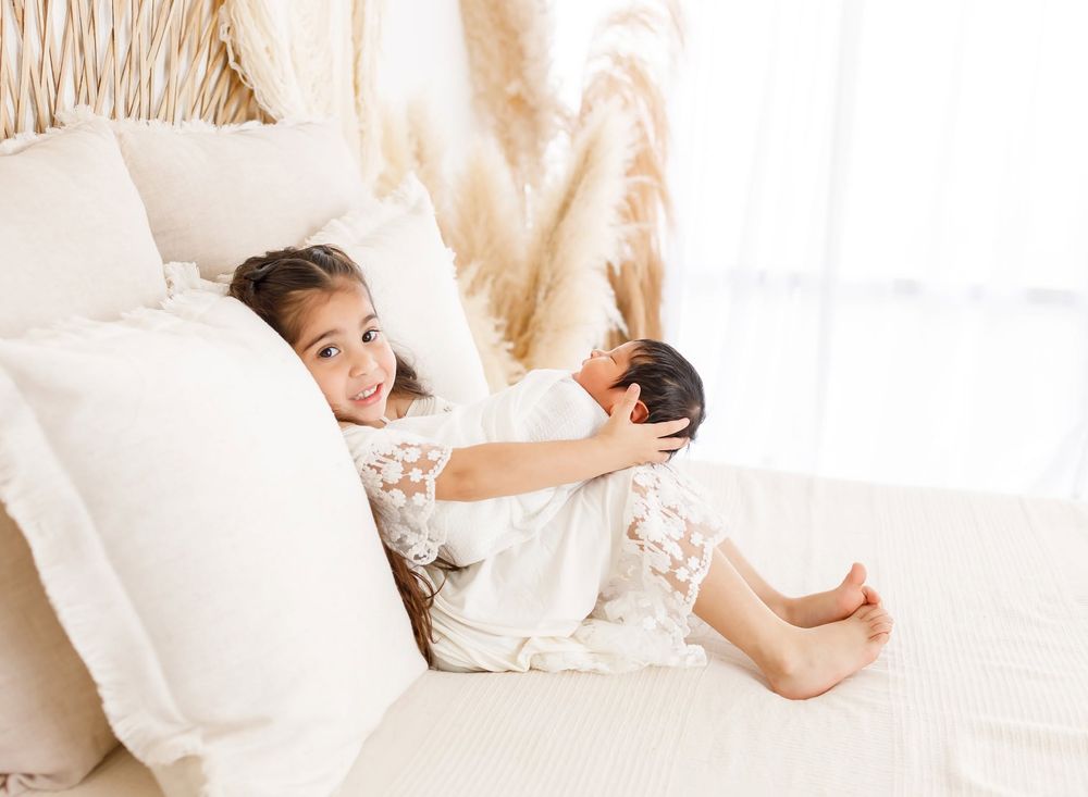 Young girl in white dress holds newborn baby on a bed with neutral boho decor.