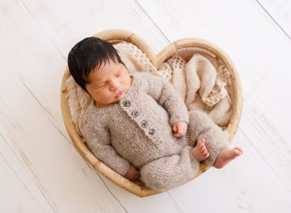 Sleeping newborn baby in knit onesie posed in heart-shaped wicker basket on white wood floor.