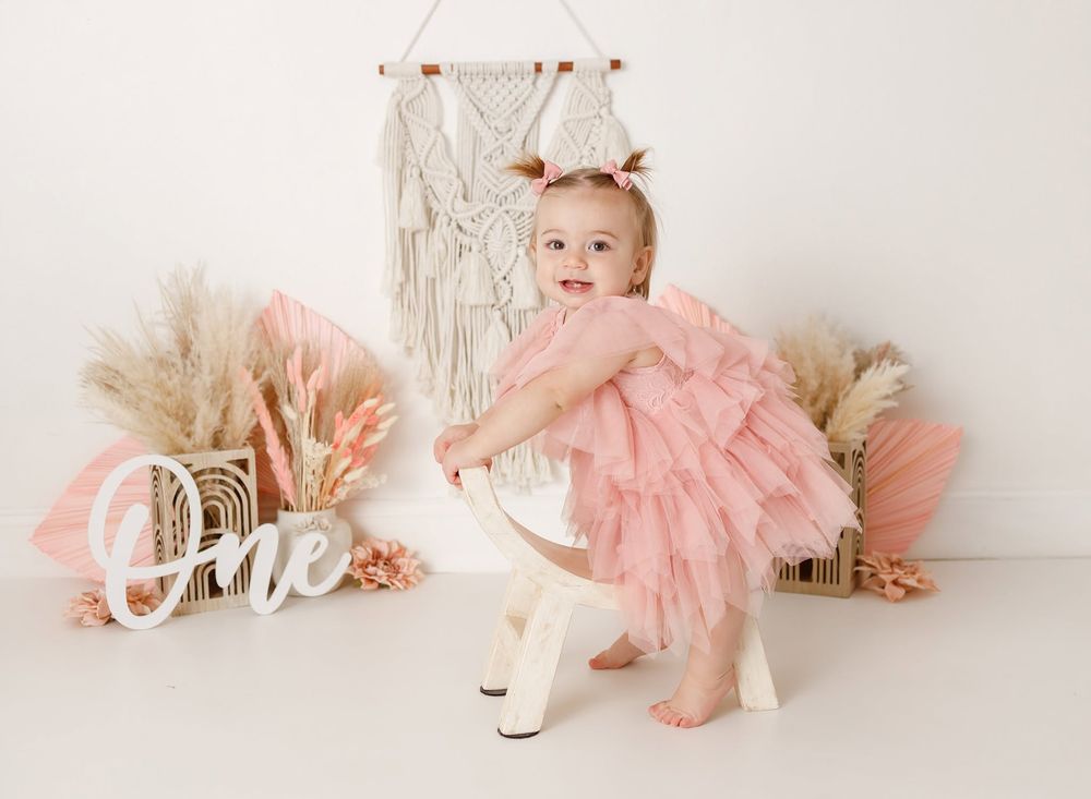 Baby girl in pink tutu dress celebrates first birthday with boho decorations and 'One' sign.