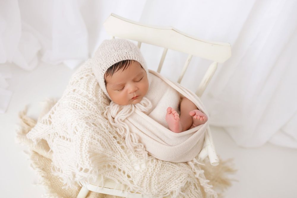 Sleeping newborn baby in cream knit outfit and bonnet, nestled in a white wooden chair with lace blanket.
