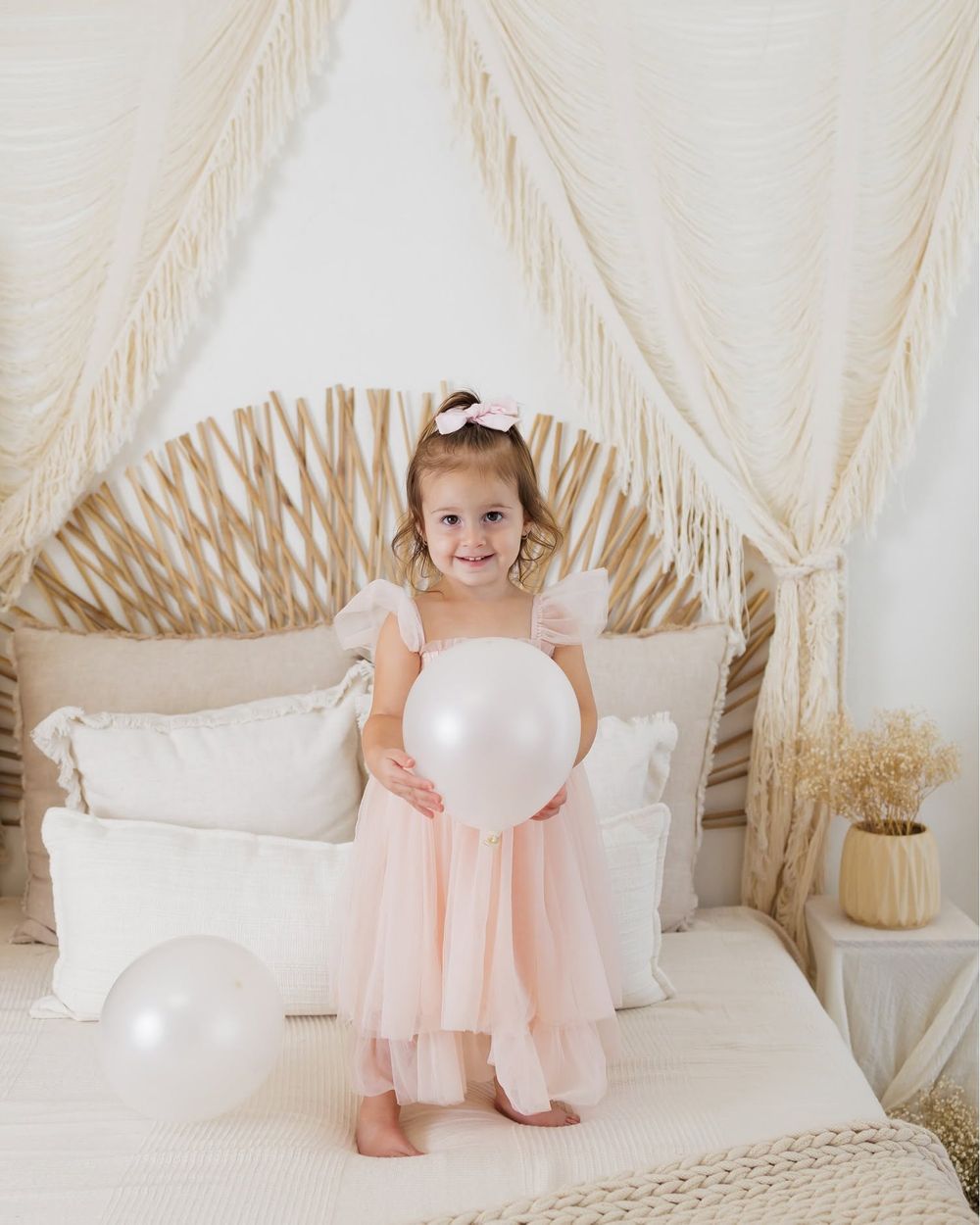 A young girl in a pink tulle dress holds a white balloon on a boho bed with macrame canopy decor.