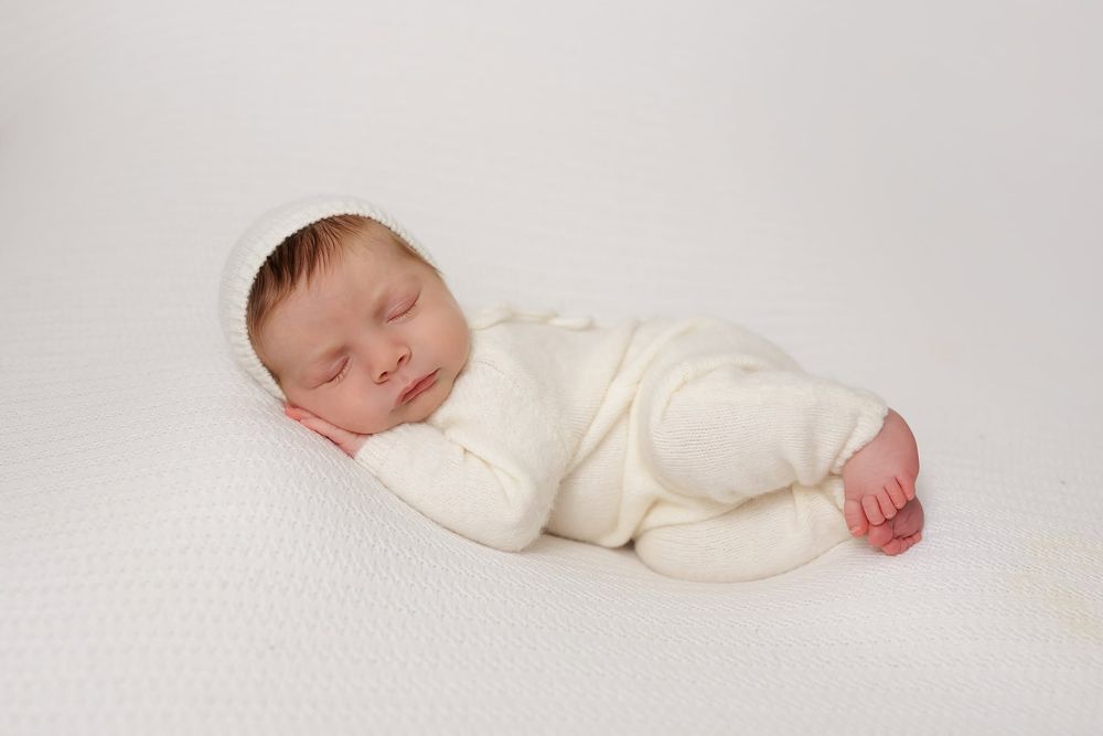 Newborn baby sleeping peacefully in white outfit and bonnet on white background.