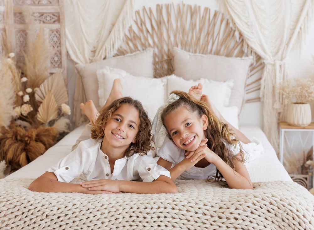 Two smiling children lying on a boho-style bed with white bedding and dried pampas grass decor.