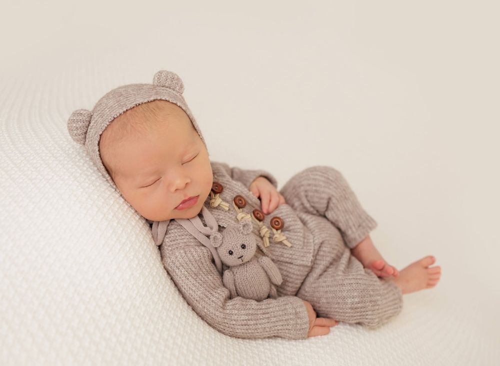 Sleeping newborn baby in gray knit bear outfit holding a tiny teddy bear on a white background.