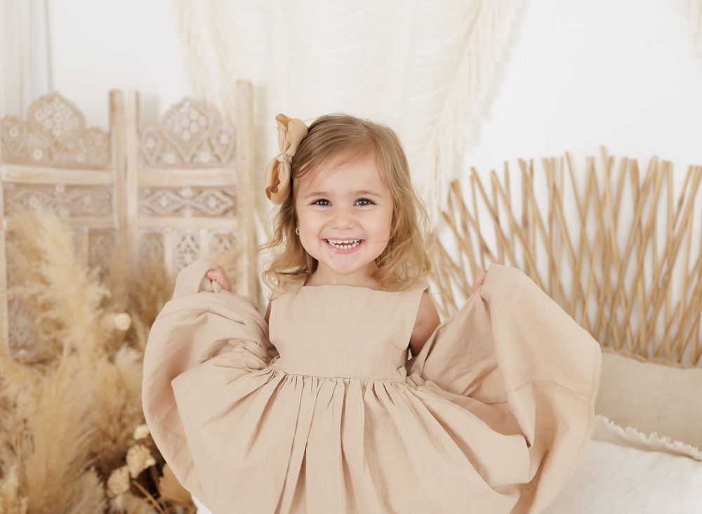 Smiling toddler girl in a tan linen dress with bow hair clip poses in a boho-styled photo studio.