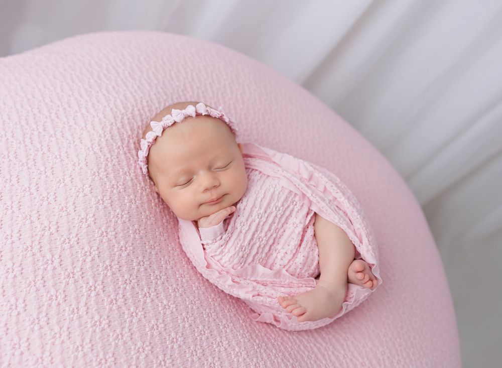 Newborn baby girl swaddled in pink wrap with floral headband, sleeping peacefully on a pink round cushion.
