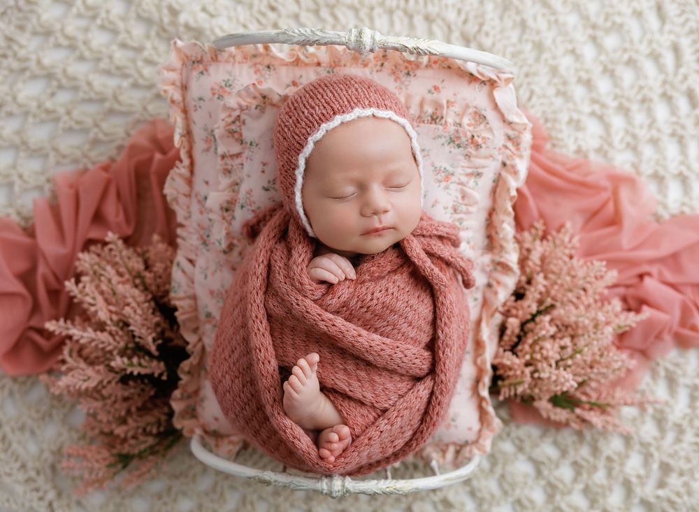 Newborn baby wrapped in pink knit swaddle with bonnet, sleeping peacefully in white bucket with floral decor.