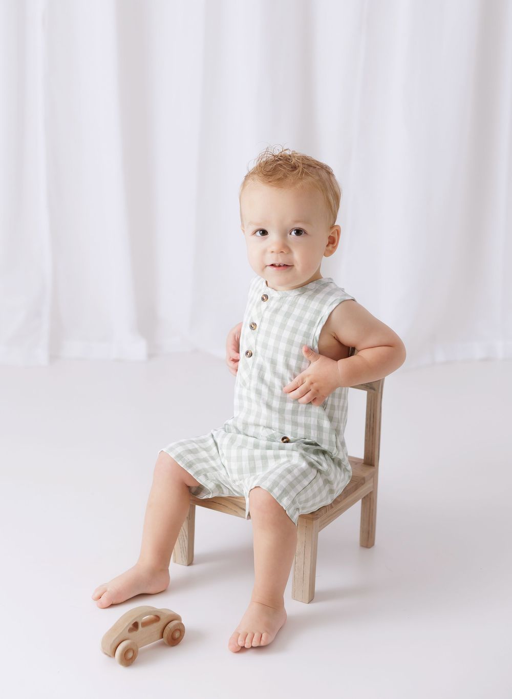 Baby boy in sage green gingham romper sitting on small wooden chair with toy car, white backdrop.