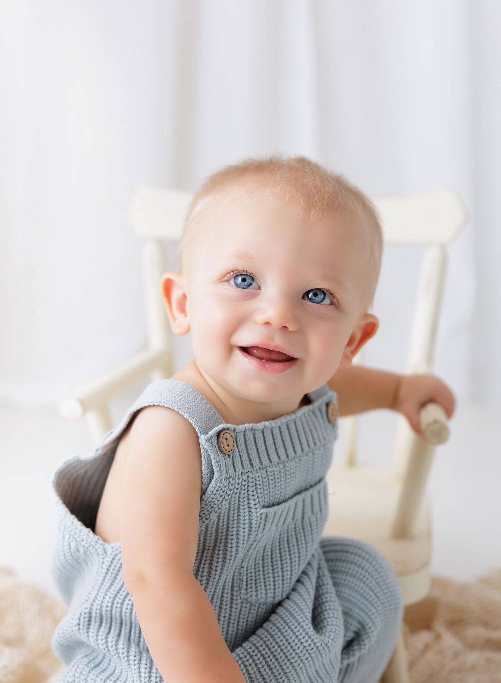 Smiling baby with blue eyes wearing a gray knit romper sits in a small white chair in a bright studio.