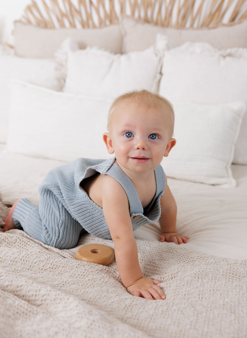 Baby in blue knit romper crawling on a white textured bed with pillows, holding a wooden toy ring.