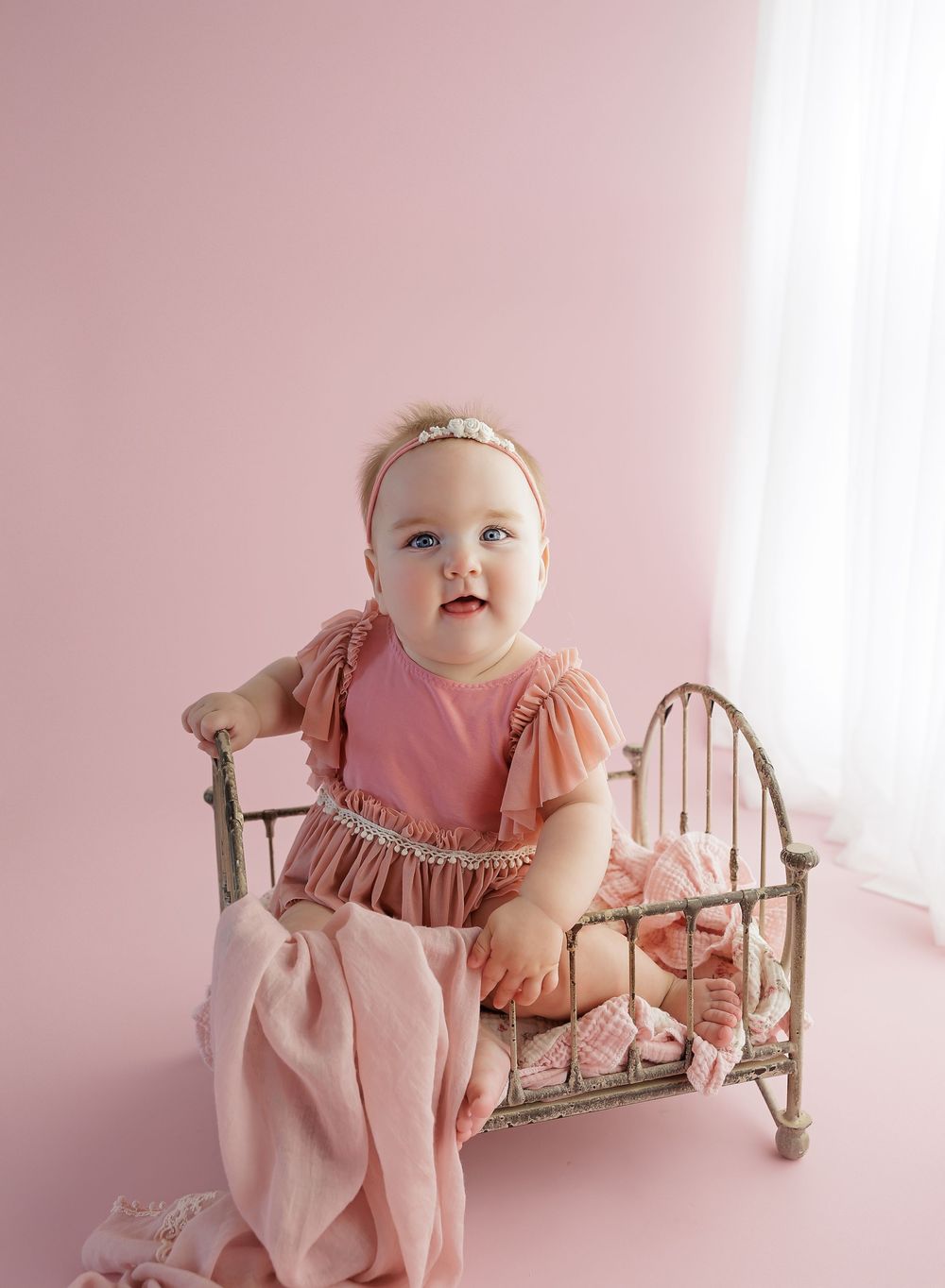 Smiling baby girl in pink romper sitting in a vintage miniature metal crib against a soft pink studio background.