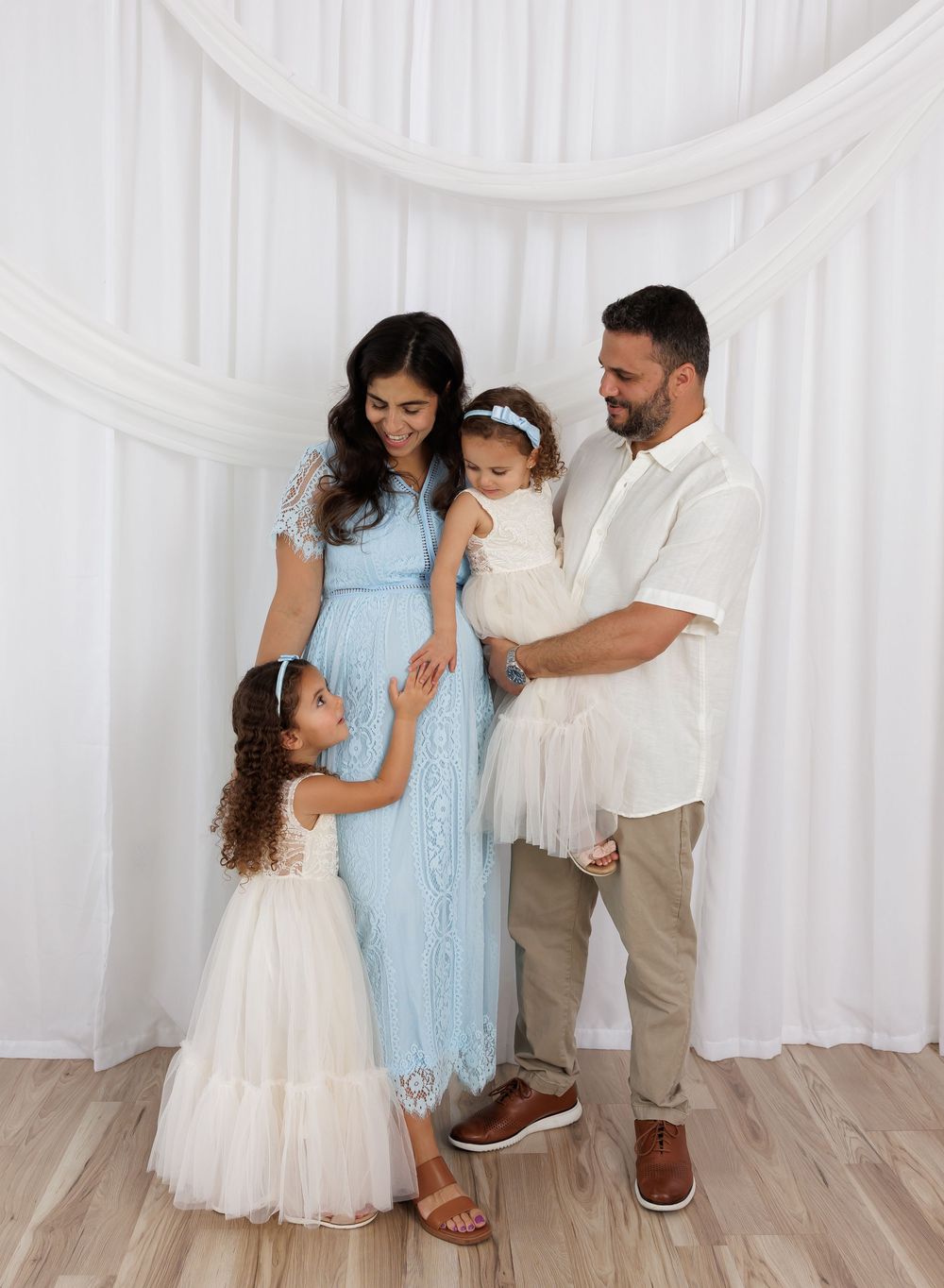 Family of four posing together, mother in blue lace maternity dress, father holding toddler, young daughter touching mom's belly.