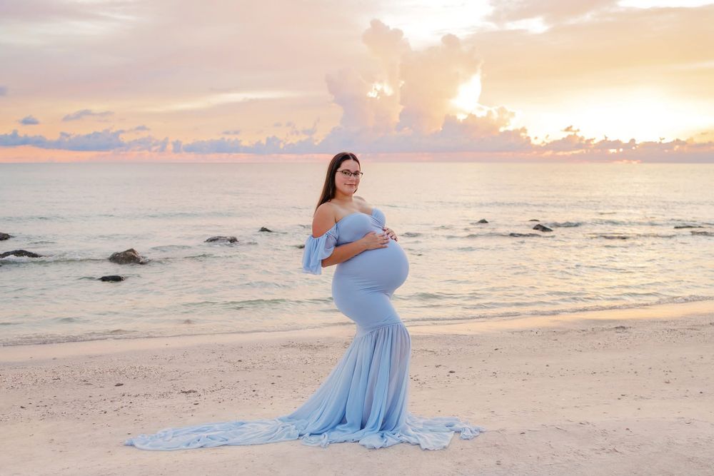 Pregnant woman in light blue gown posing on beach at sunset, cradling baby bump with ocean waves behind her.