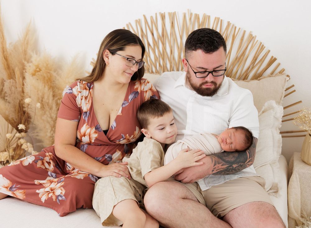 Family of four posing for a newborn photo session, parents with toddler holding sleeping baby on a white couch.