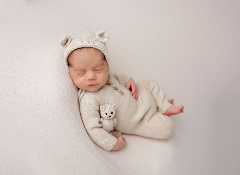 Newborn baby sleeping peacefully in cream knit bear outfit and bonnet, holding a tiny teddy bear on white background.