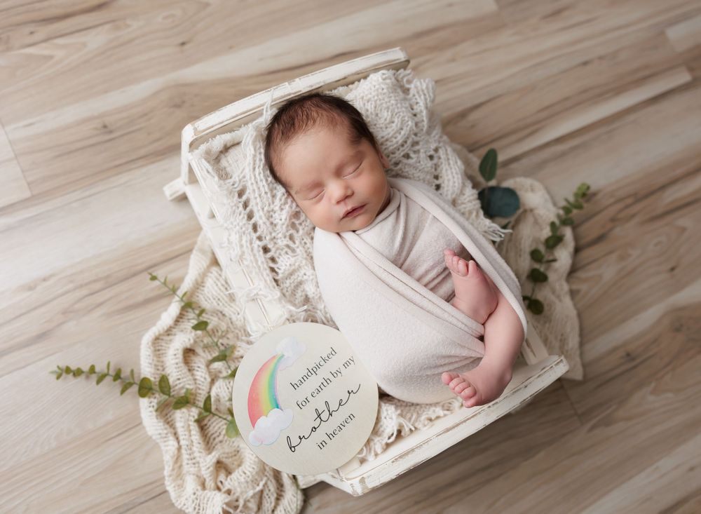 Newborn baby swaddled in white wrap sleeping in a white wooden box with lace blanket and a rainbow milestone card.