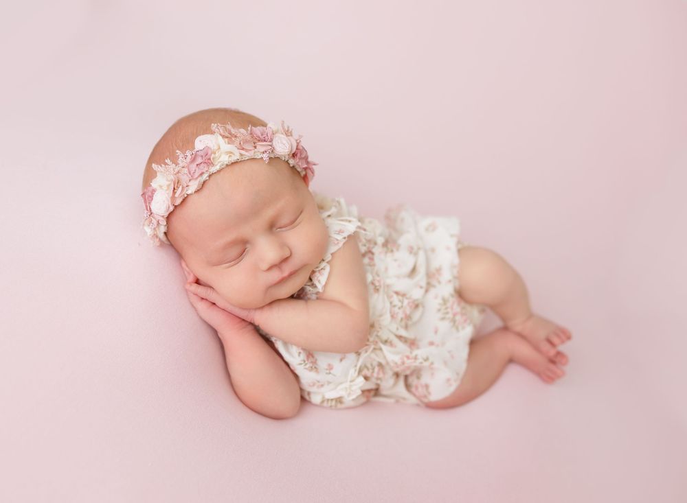 Sleeping newborn baby girl in white lace romper and pink floral headband on soft pink backdrop.