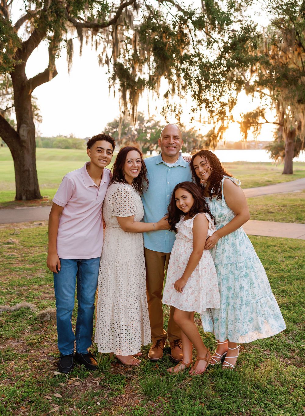 Family of five smiling together outdoors at golden hour under Spanish moss-draped trees near a lake.
