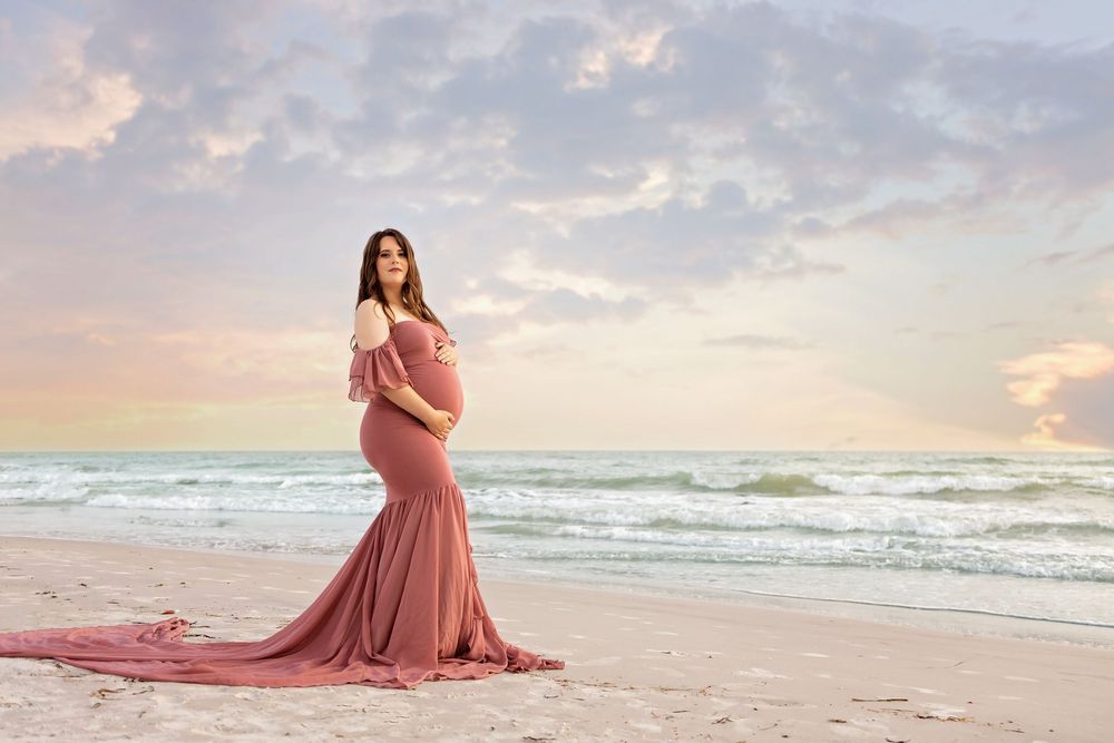 Pregnant woman in pink mermaid gown poses on beach at sunset with ocean waves and pastel sky.