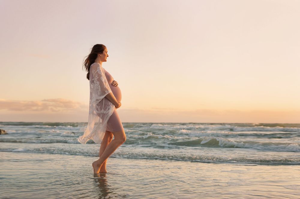 Pregnant woman in white dress walking barefoot on beach at sunset, cradling her baby bump by ocean waves.