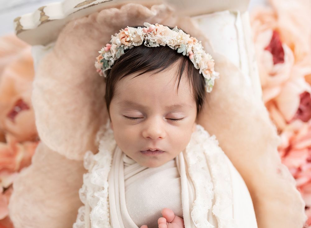 Sleeping newborn baby girl wearing a delicate floral crown, wrapped in cream lace, surrounded by peach roses.
