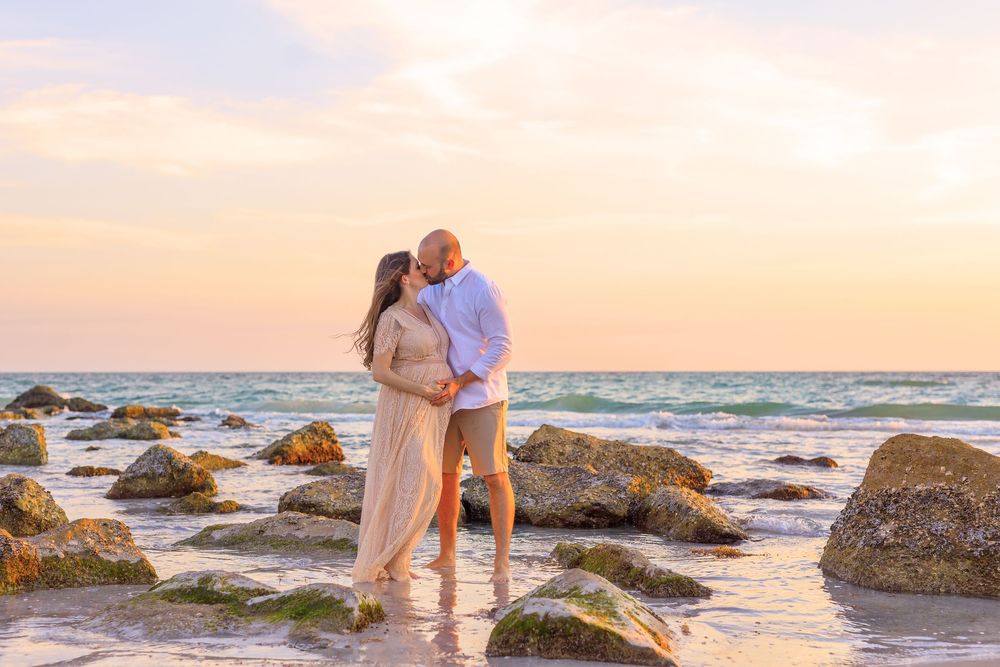 Couple embracing romantically on a rocky beach at sunset with pastel skies and ocean waves in the background.