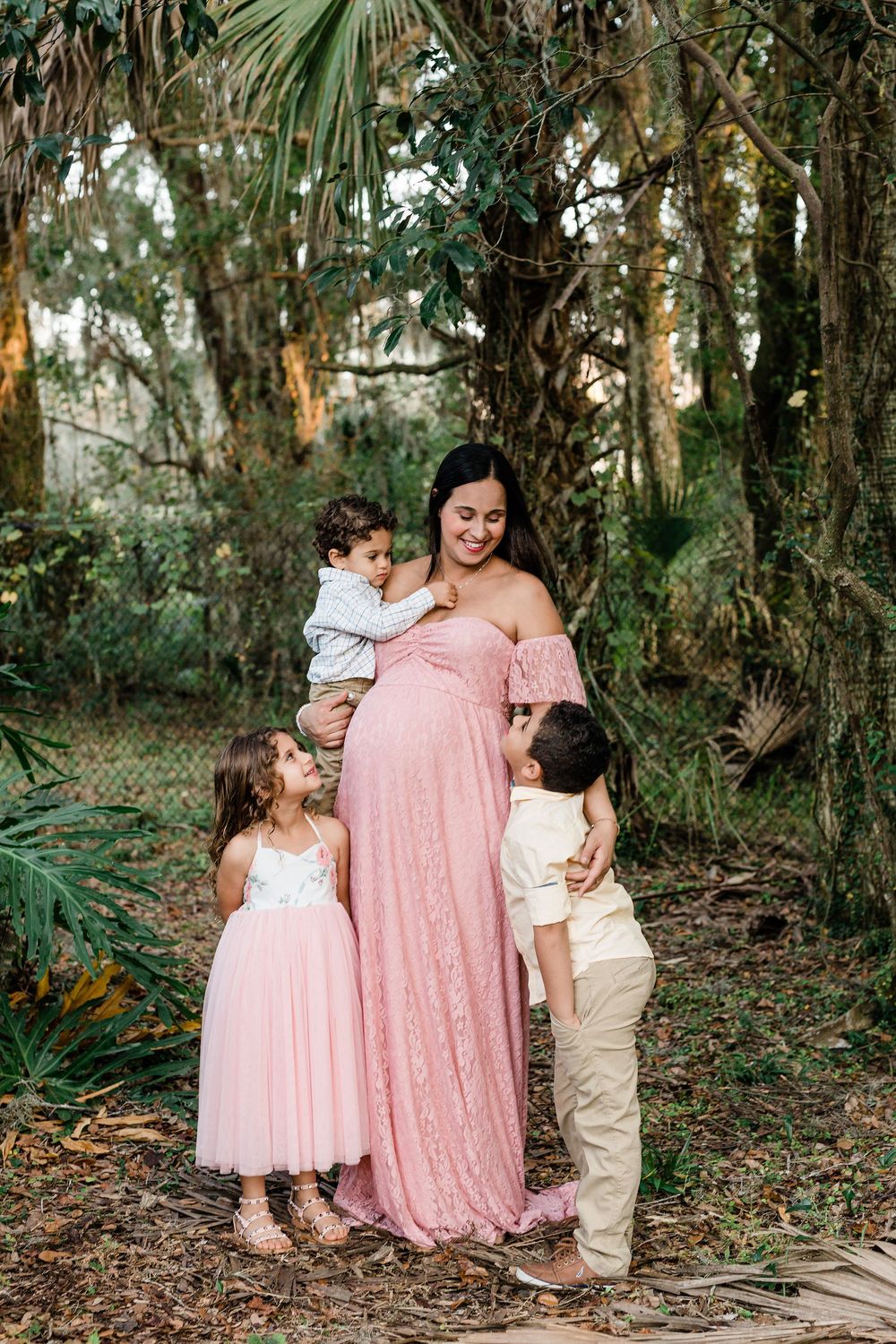 Pregnant woman in pink lace gown holds toddler while two young children embrace her baby bump outdoors.
