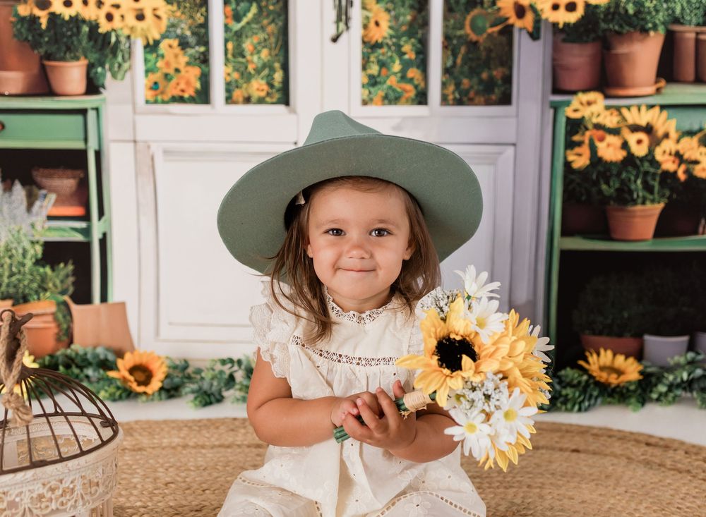 A smiling little girl wearing a green hat holds a sunflower bouquet in a floral garden-themed photo backdrop.