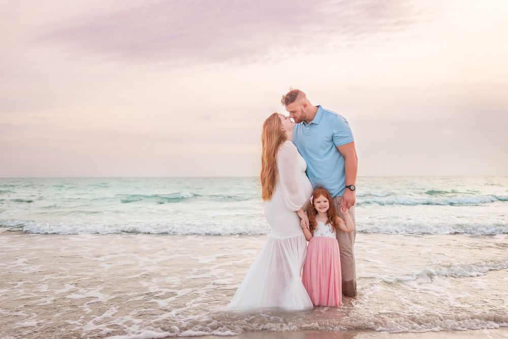 Family beach portrait at sunset with parents sharing a kiss while young daughter in pink dress stands between them.