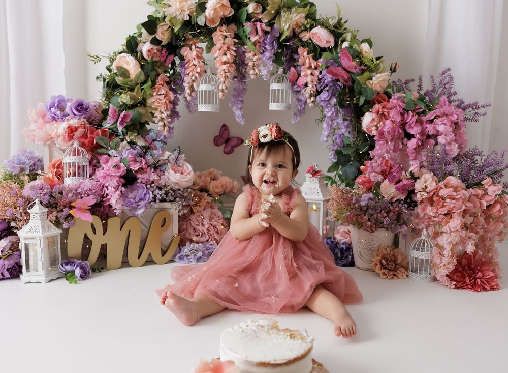 Baby girl in pink dress sits smiling at her first birthday cake smash with floral arch backdrop.