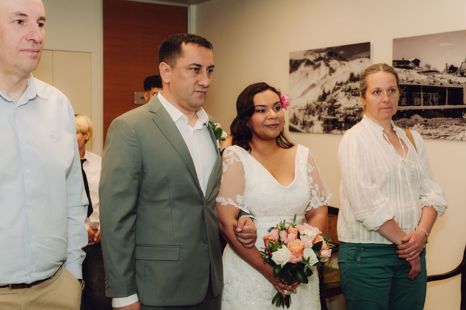 Bride holding pink bouquet walks with groom in gray suit during indoor wedding ceremony.