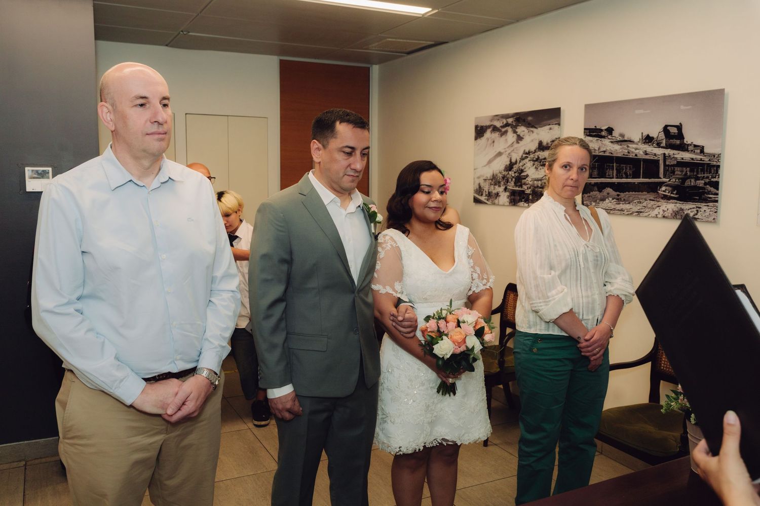 Wedding party of four stands together indoors, bride holding pink bouquet in white dress.
