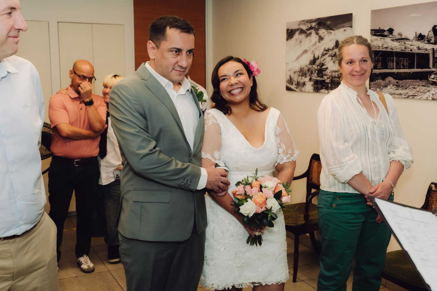 Smiling bride holding pink bouquet stands with groom in gray suit during indoor wedding ceremony.