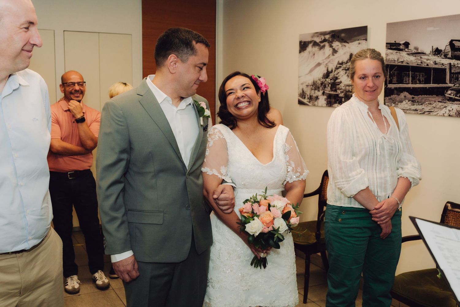 Happy bride with pink flower in hair holds bouquet beside groom in gray suit at wedding.