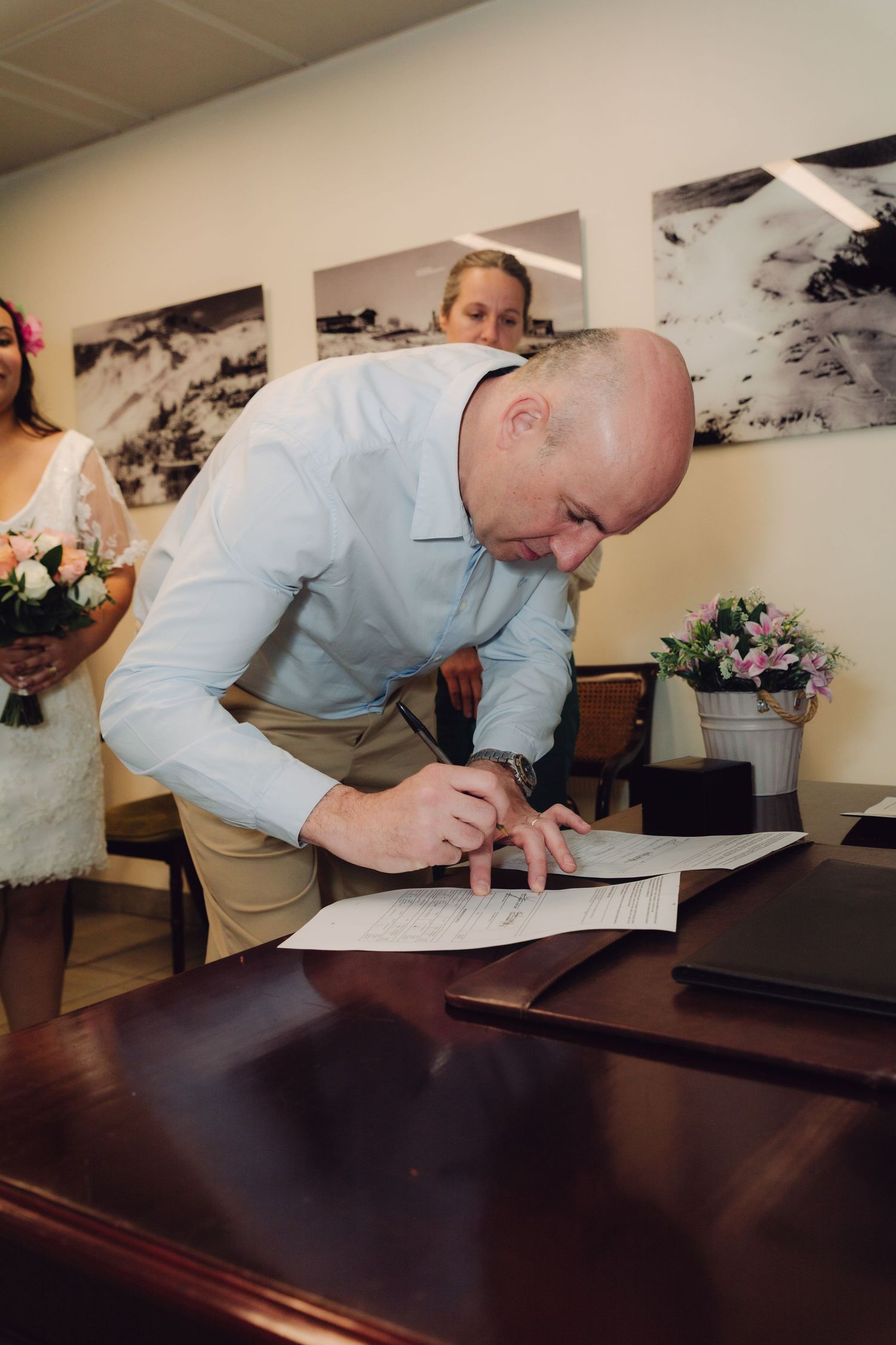Bald man signs official wedding documents at wooden desk during civil marriage ceremony.