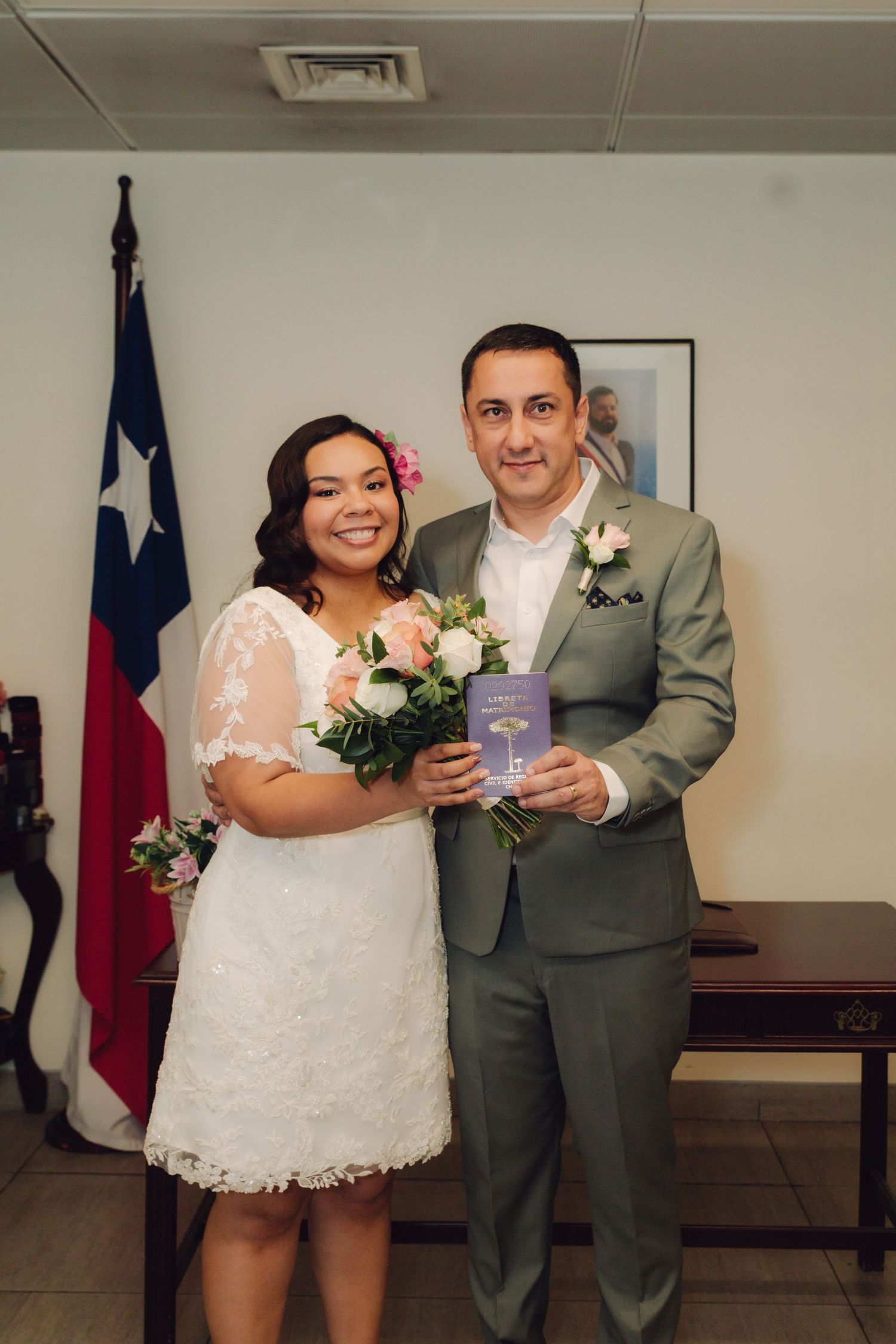 Newlyweds pose with marriage certificate in front of Chilean flag after civil wedding ceremony.